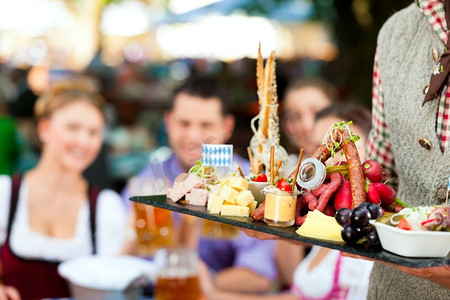 巴伐利亚州摄影照片_在德国巴伐利亚州的啤酒花园-朋友Tracht、Dirndl和一张桌子上，摆着啤酒和小吃