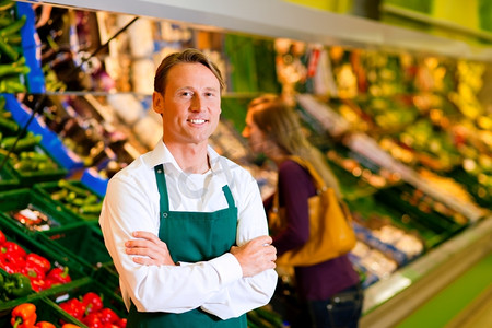 杂货食品摄影照片_在蔬菜货架的超级市场的店员;在背景中一个选择蔬菜的妇女