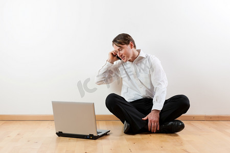 一名男子盘腿坐在公寓的木地板上，手里拿着笔记本电脑，正在打电话