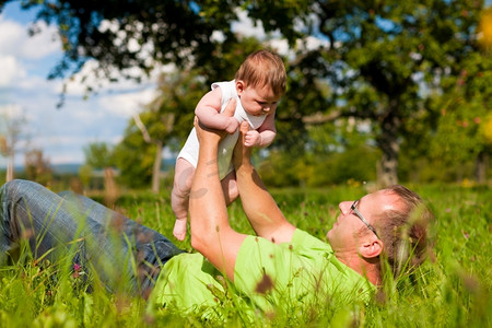 绿色花草草地摄影照片_在一个阳光明媚的日子里，父亲带着他的孩子在绿草如茵、花草丛生的草地上玩耍。