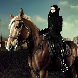 马上开战摄影照片_优雅的女人穿着黑色大衣骑在棕色的马上