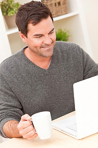 微笑的人在他的三十多岁在家里使用他的笔记本电脑drinkng茶或咖啡