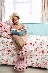 年轻女孩(5-6岁)穿着兔子装和怪物拖鞋坐在床上看书