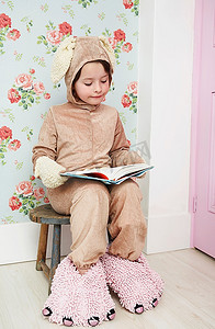 年轻女孩(5-6岁)穿着兔子装和怪物拖鞋坐在凳子上看书
