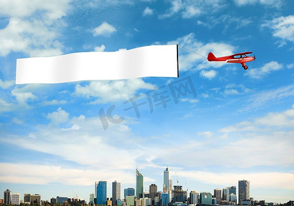 城市上空升起一架飞机，挂着一面空白的旗帜