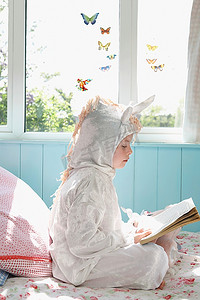 年轻女孩(5-6岁)穿着独角兽服装坐在床上看书