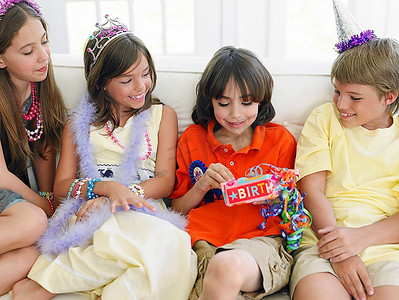 礼物长摄影照片_四个孩子(7-12岁)在生日派对上坐在沙发上，一份开幕礼物
