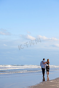 沐光而行摄影照片_一对年轻的男女浪漫情侣在蔚蓝天空的海滩上相拥而行