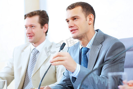 图为两个商人坐在会议桌旁,用麦克风讲话合照人物特写