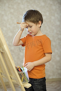 一个小男孩用一支铅笔把手举到脸上，站在一块画布前面，手里拿着一罐彩色铅笔