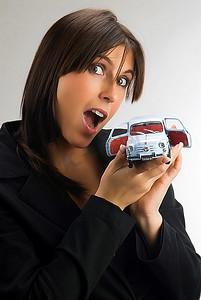 一位年轻可爱的黑发女子惊讶地看着镜头，展示着一辆意大利汽车菲亚特600的模型