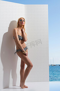 夏天的美丽女孩穿着比基尼，在白墙附近摆姿势，臀部有纹身。大海，蓝天和一些船作为背景