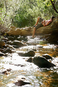 小溪边摄影照片_年轻男子躺在小溪边的木头上