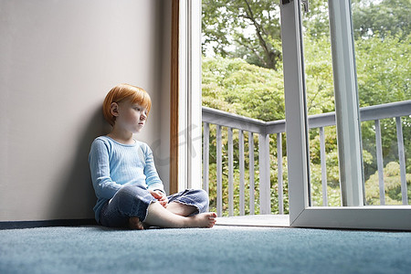 年轻女孩（5-6）盘腿坐在地毯上，通过阳台门看