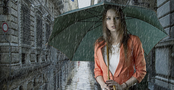 美丽的年轻女子撑着绿色的雨伞，在雨中飘落在一条古老的时尚街道上