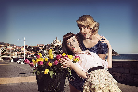 时尚花卉画摄影照片_有葡萄酒样式的两个时尚妇女的画象在滑稽的姿势的在自行车的篮子附近有一些五颜六色的花在葡萄酒颜色的海附近