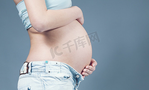 腹部的特写，双手放在上面。怀孕的白人妇女穿着浅蓝色上衣和牛仔裤。孕期收藏。
