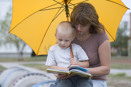 年轻的男孩和女人坐在黄色的伞下看书