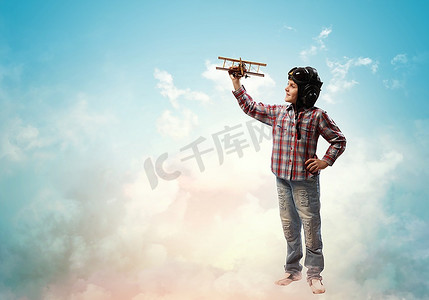 玩玩具男孩摄影照片_戴着飞行员头盔的小男孩在云的背景下玩玩具飞机