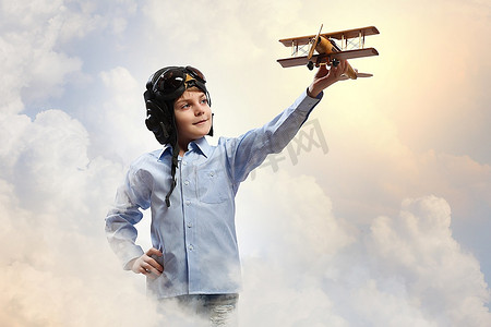玩玩具的男孩摄影照片_戴着飞行员头盔的小男孩在云的背景下玩玩具飞机