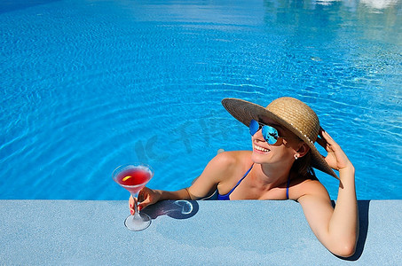 泳游泳摄影照片_戴着帽子的女人在泳池边放松，喝着国际化的鸡尾酒