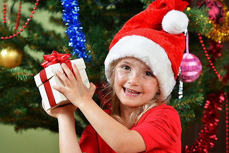 圣诞节圣诞帽摄影照片_圣诞树附近戴着圣诞帽的漂亮小女孩