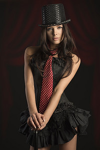 性感的黑发女子在歌舞表演中穿着深色紧身胸衣，戴着帽子和红色领带。在黑色背景上用棍子摆姿势