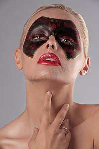 黑红电竞摄影照片_美丽的金发女孩肖像，脸上涂着黑红相间的狂欢节面具，涂着红色口红