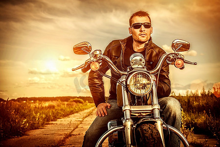 一名骑自行车的男子穿着皮夹克，戴着墨镜，坐在摩托车上看着日落。