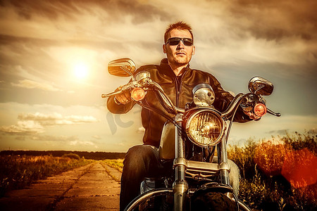 骑摩托的男人摄影照片_一名骑自行车的男子穿着皮夹克，戴着墨镜，坐在摩托车上看着日落。