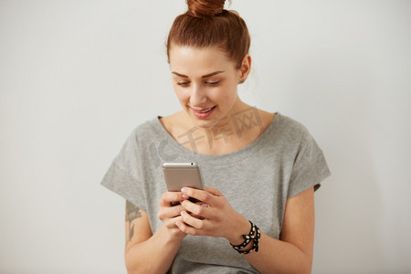 手机版电商首页摄影照片_一个微笑的年轻女子使用手机的肖像。美丽的快乐女孩聊天和短信在灰色背景的手机。年轻女子在智能手机上阅读短信