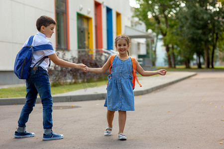 学习摄影照片_一个笑容满面的小女孩和一个背着背包的弟弟手牵着手，玩耍着，在教学楼附近玩得很开心