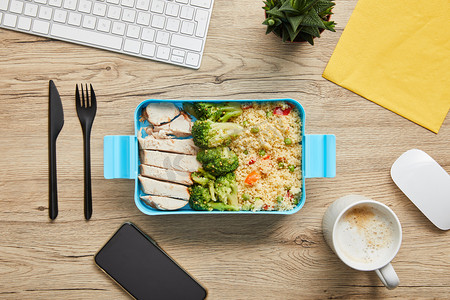 午餐盒的顶视图与鸡肉，花椰菜和利索托在桌子上与电脑键盘和智能手机