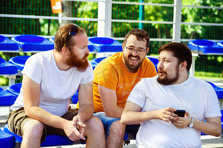 Three male friends are watching mobile phone video and laughing sitting at summer day at stadium background.