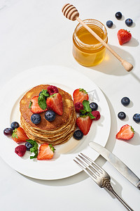 菜煎饼摄影照片_顶部的美味煎饼与蜂蜜，蓝莓和草莓盘在叉子附近，刀在大理石白色表面