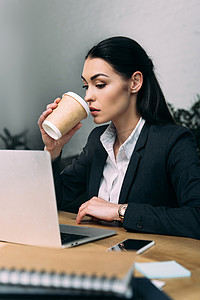 在办公室的笔记本电脑在工作场所的女商人的侧面视图与一次性杯咖啡