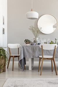 白色椅子和桌子上方的灯, 在灰色的餐厅内部, 有植物和圆形镜子。真实照片