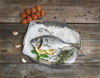 Roasted dorado or sea bream fish with vegetables, herbs and spices on silver tray over rustic wood backdrop, top view