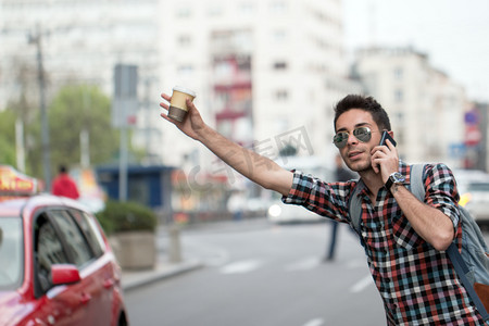 Young man on the street of big city , tries to catch taxi. Carry backpack on one shoulder, street, peaople and cars in background. Casual dressed.
