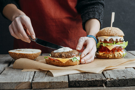桌面制作摄影照片_在围裙上的女孩裁剪视图制作美味自制芝士汉堡在木制桌面上烘烤纸