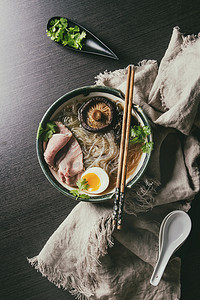 日本烹饪摄影照片_传统的日本面条汤, 蘑菇, 鸡蛋, 切片牛肉和蔬菜, 在陶瓷碗中供应木筷和白色勺子, 在黑暗的背景布。平躺, 空间.