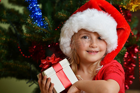 圣诞帽绿色摄影照片_圣诞树附近戴着圣诞帽的漂亮小女孩