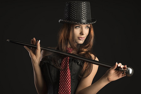 美丽的黑发女子穿着像性感的卡巴莱表演者，戴着帽子，穿着紧身胸衣，打着领带，在黑色背景下玩棍子