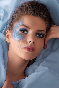 一位漂亮的年轻女子涂着蓝色闪闪发光的化妆品，躺在布片上的面部照片