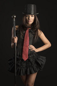 漂亮的黑发女子穿着像歌舞演员一样的衣服，黑色紧身胸衣，红色领带，黑色背景下手拿棍子的帽子