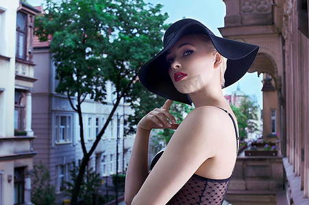 年轻的金发时尚女郎在时尚照中戴着优雅的黑色帽子和胸罩站在窗外，背景是古老的宫殿