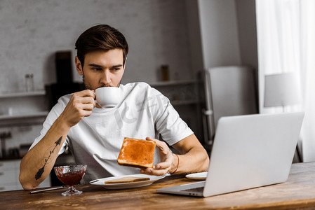 电子电器摄影照片_英俊的年轻自由职业者吃烤面包与果酱和咖啡, 看着笔记本电脑屏幕在家里