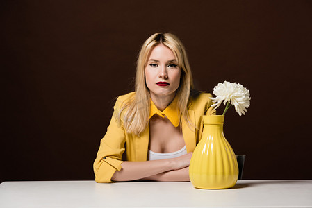 美丽的时尚金发女孩看着相机, 而坐在餐桌上, 白色的花朵在棕色的黄色花瓶