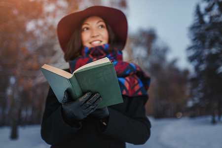 雪摄影照片_红色帽子持有和阅读诗歌书在冬季森林背景的年轻女子