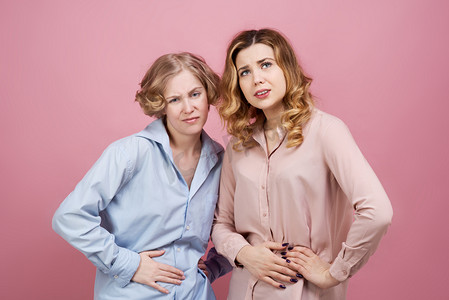 两个年轻的女人用痛苦的表情抱着他们的肚子。工作室肖像粉红色背景。胃病、月经痛、消化不良的概念.